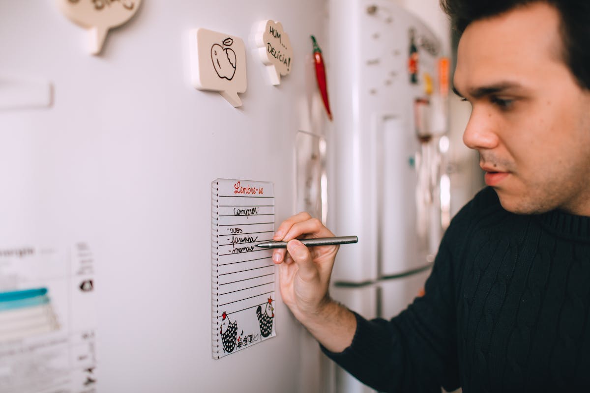 A person writing a reminder list on a note attached to a refrigerator.